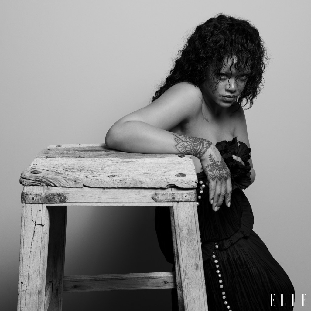 A Black and white portrait of a woman with curly hair and tattoos, sitting on a wooden bench with a pensive expression.