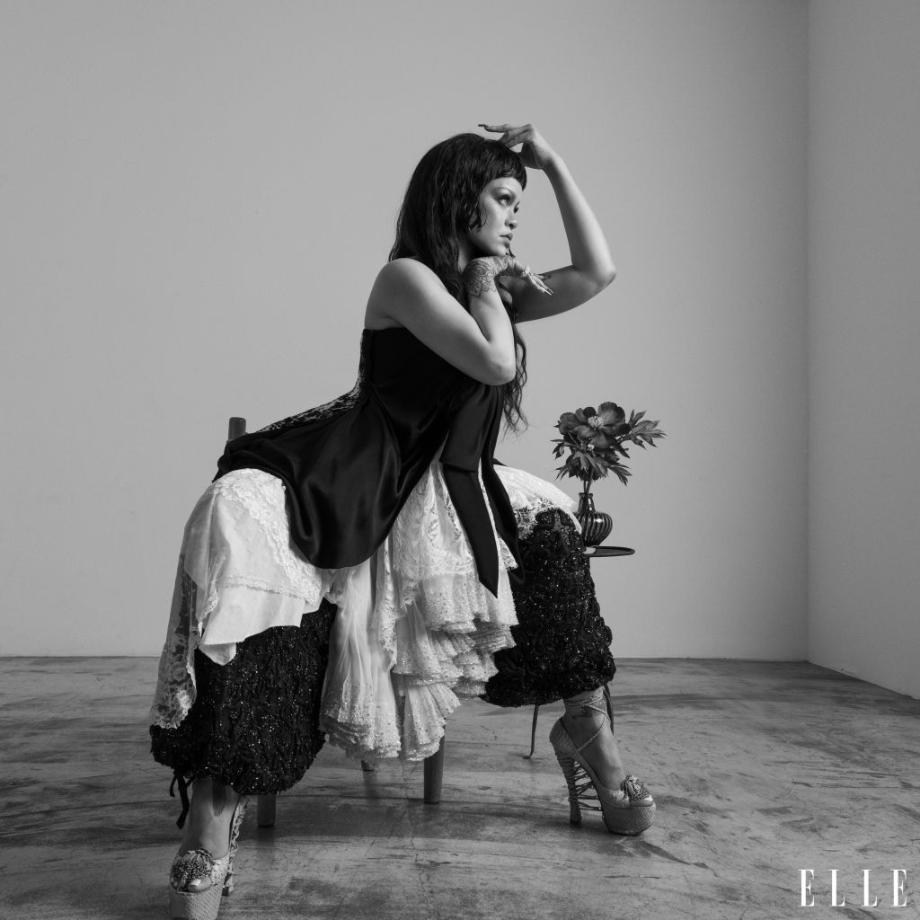 A woman in a black dress and lace skirt stands with her arms raised, holding a bouquet of flowers in a studio setting.