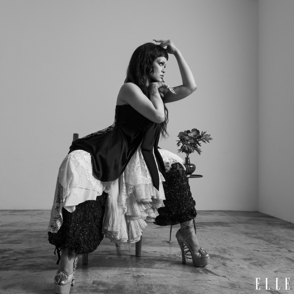 A woman in a black dress and lace skirt stands with her arms raised, holding a bouquet of flowers in a studio setting.