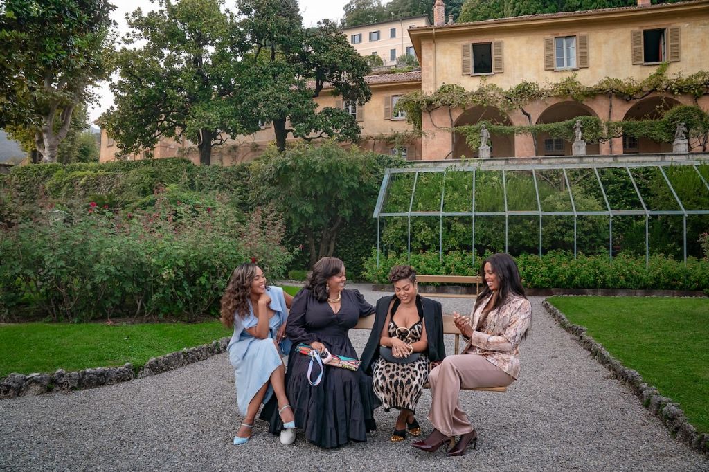 Four women in formal dresses sitting on a bench in a lush garden with a greenhouse in the background.