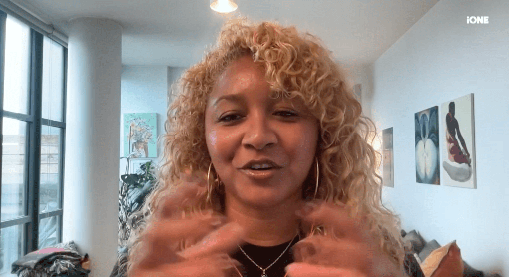 A middle-aged woman with curly blonde hair and a necklace speaking in front of a window with artwork on the walls behind her.