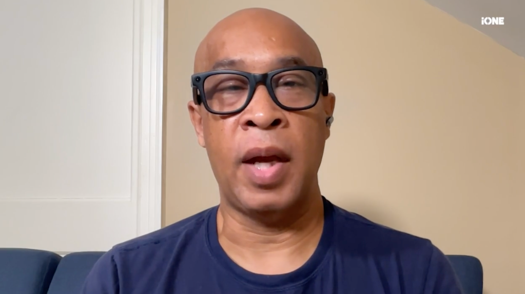 A bald man wearing glasses and a navy blue shirt looking directly at the camera with a serious expression.
