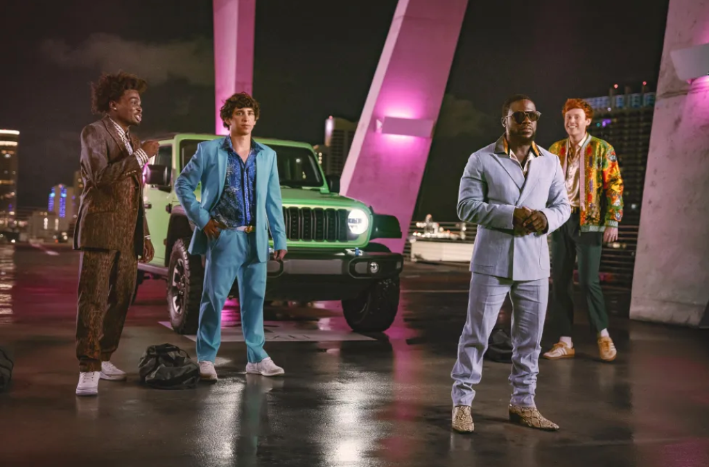 Four people standing in front of a green Jeep Wrangler at night, with neon pink and purple lighting in the background.