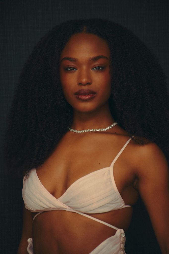 A close-up portrait of a young Black woman with long, dark curly hair wearing a white, low-cut top and a diamond choker necklace against a dark background.