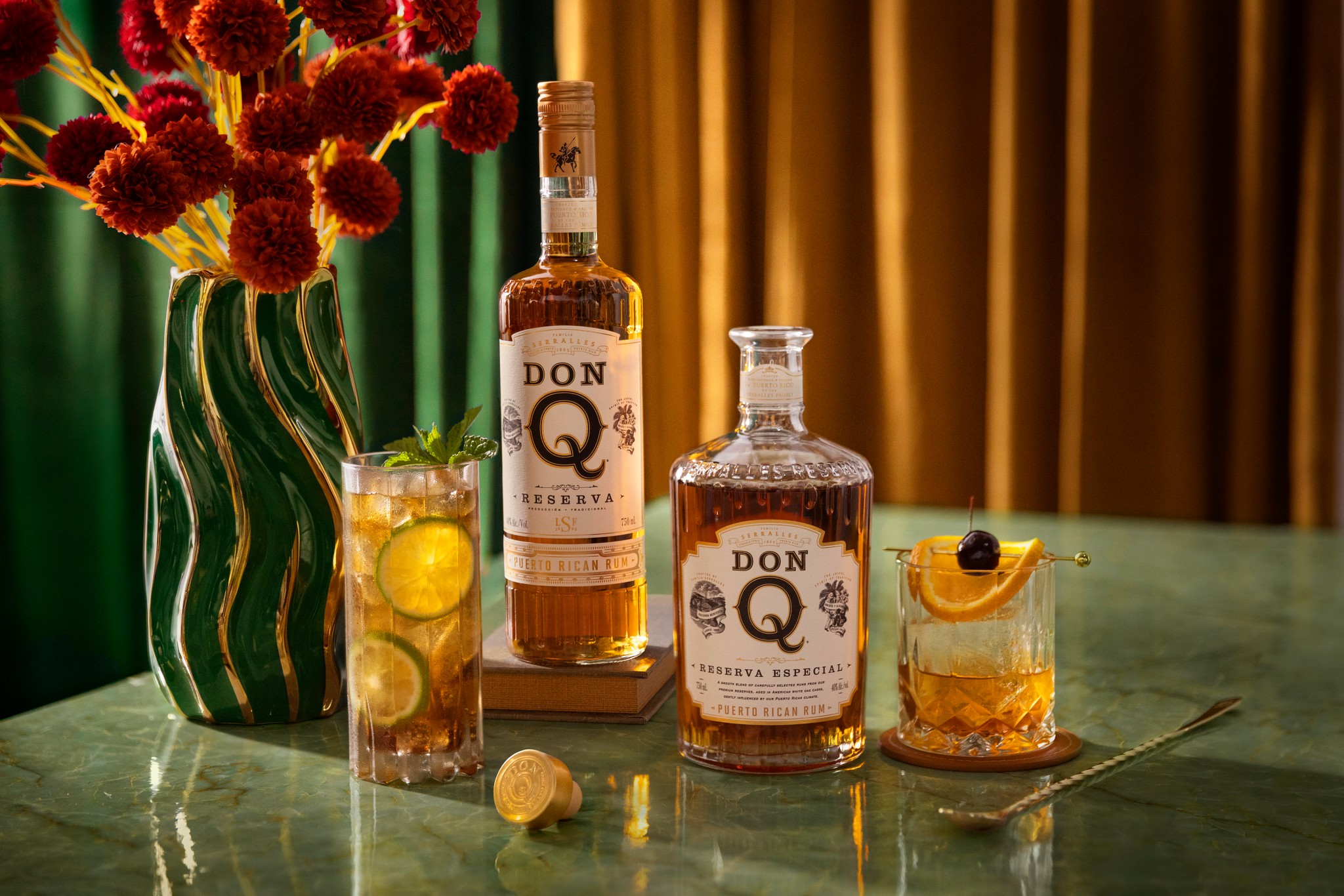 Bottles of Don Q rum, glasses of cocktails, and a vase of red flowers on a wooden table.