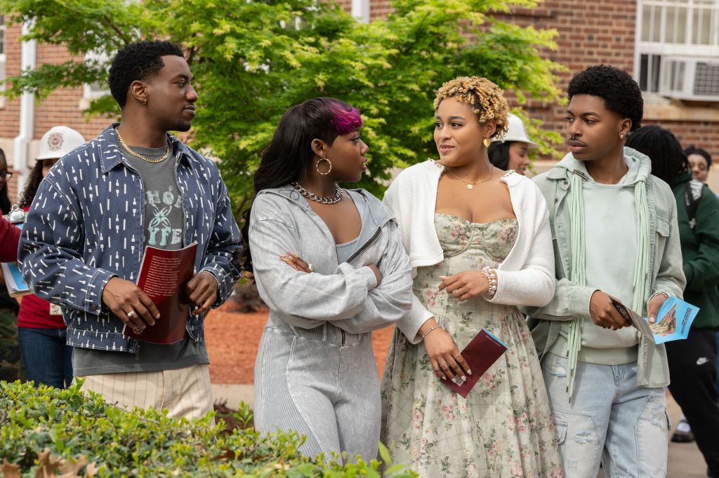 "Four young adults stand together outdoors on a college campus, dressed in casual and semi-formal clothes, holding pamphlets and talking, with trees, other people, and a brick building in the background."
