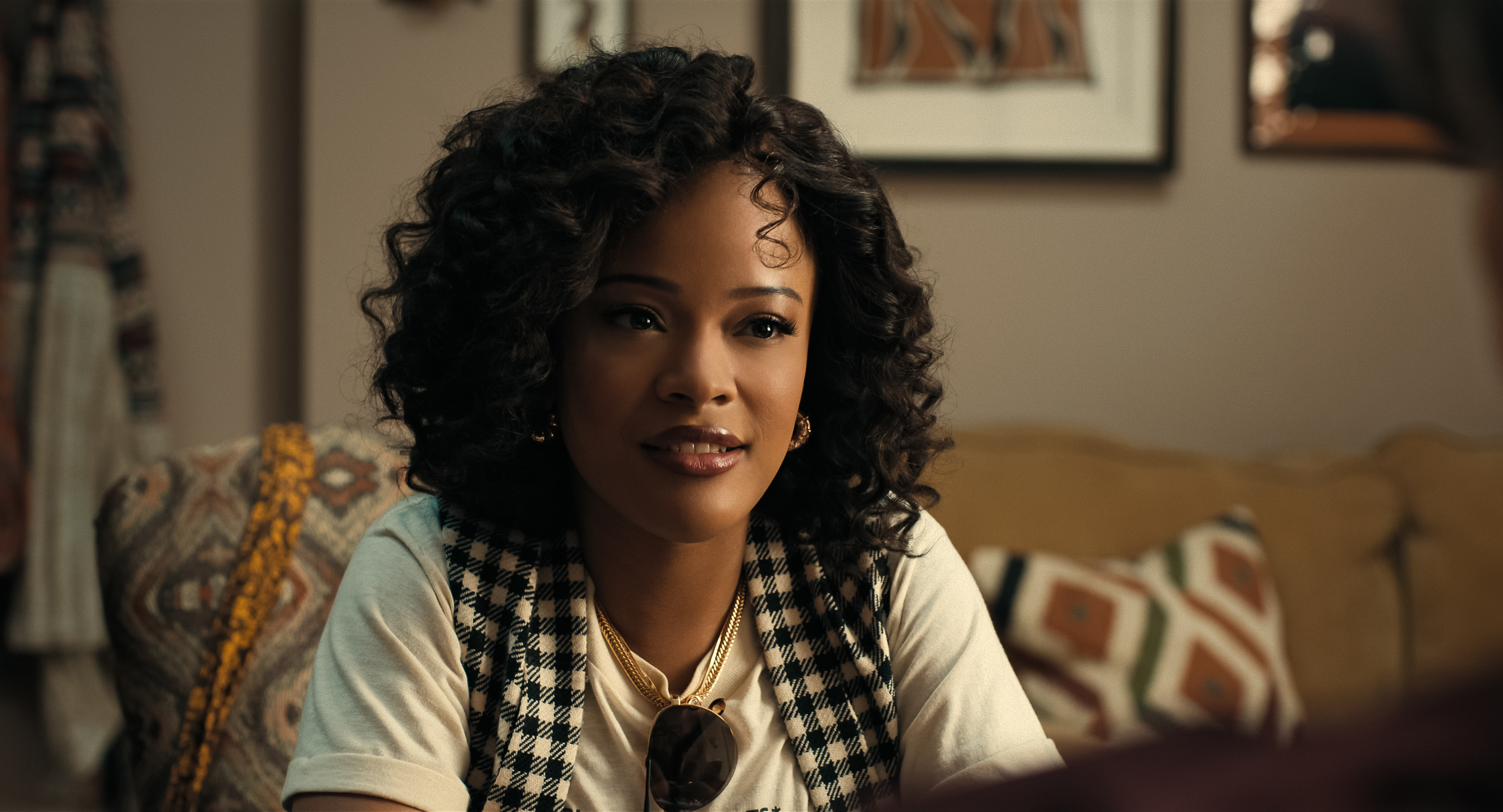 "A young woman with curly hair sits on a patterned couch in a cozy living room, wearing a checkered vest and white shirt, with framed art and cushions in the background, engaged in conversation."