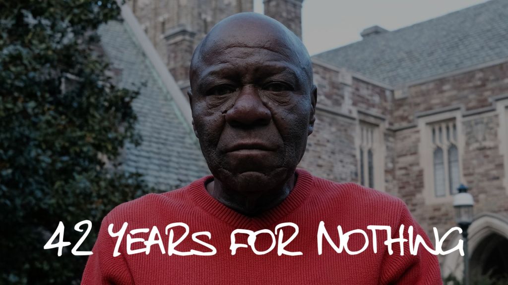 An older Black man wearing a red sweater with the text "42 YEARS FOR NOTHING" printed on it, standing in front of a stone building.