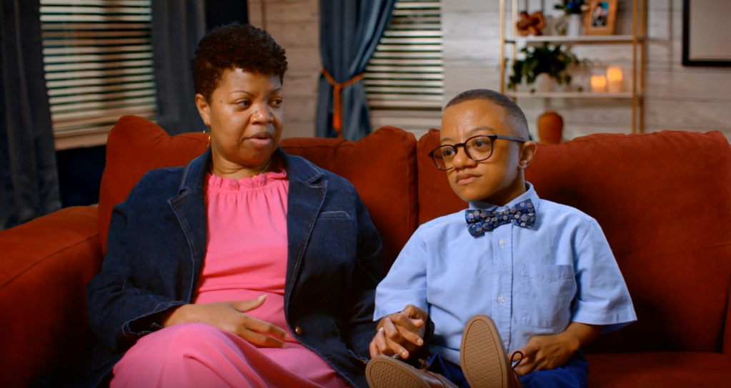 Two people, an adult woman and a young boy, sitting on a red couch in a living room setting.