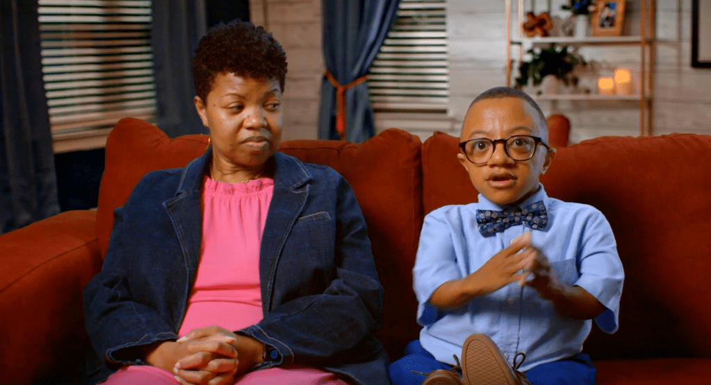 Two people, an adult woman and a young boy, sitting on a couch in a living room setting. The boy is wearing a blue shirt with a bow tie.