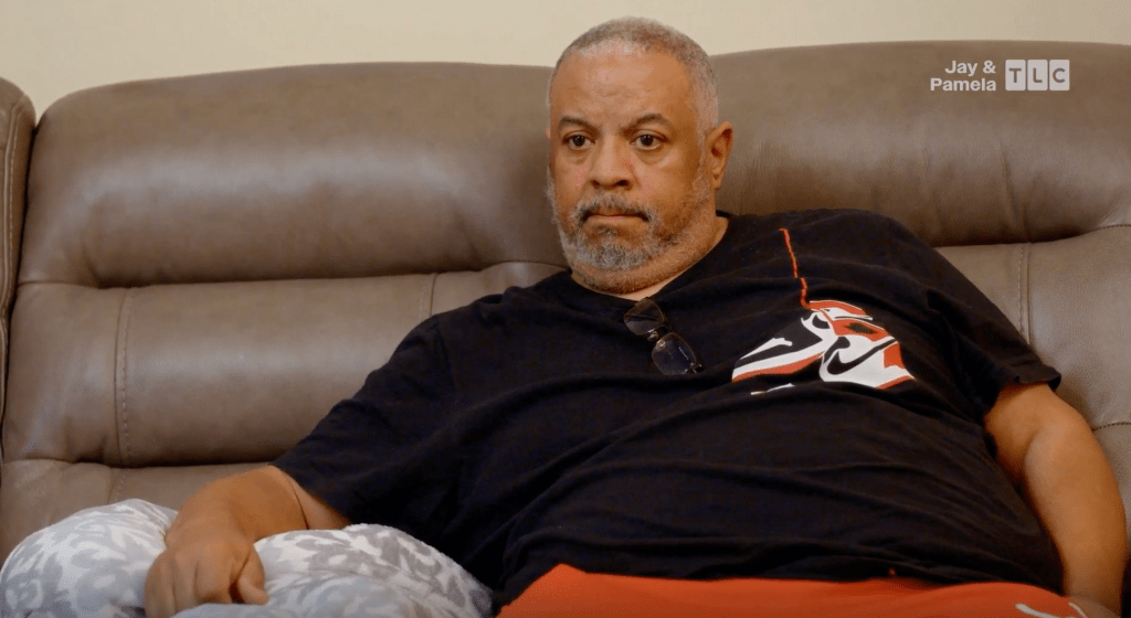 A middle-aged man with gray hair and a beard sitting on a leather couch wearing a black shirt with a Nike logo.