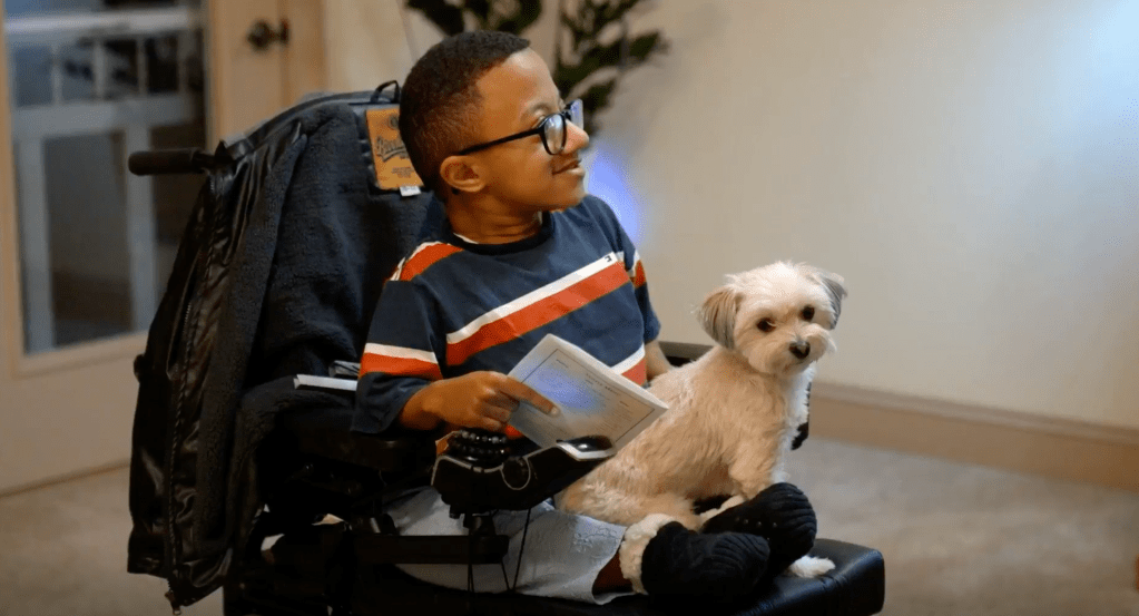 A young boy in a wheelchair holding a book and sitting next to a small white dog.