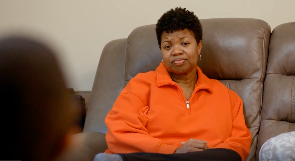 A middle-aged Black woman wearing an orange jacket sitting on a gray leather sofa.