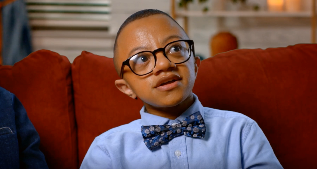 A young boy wearing glasses and a bow tie, sitting on a red couch and looking thoughtfully at the camera.