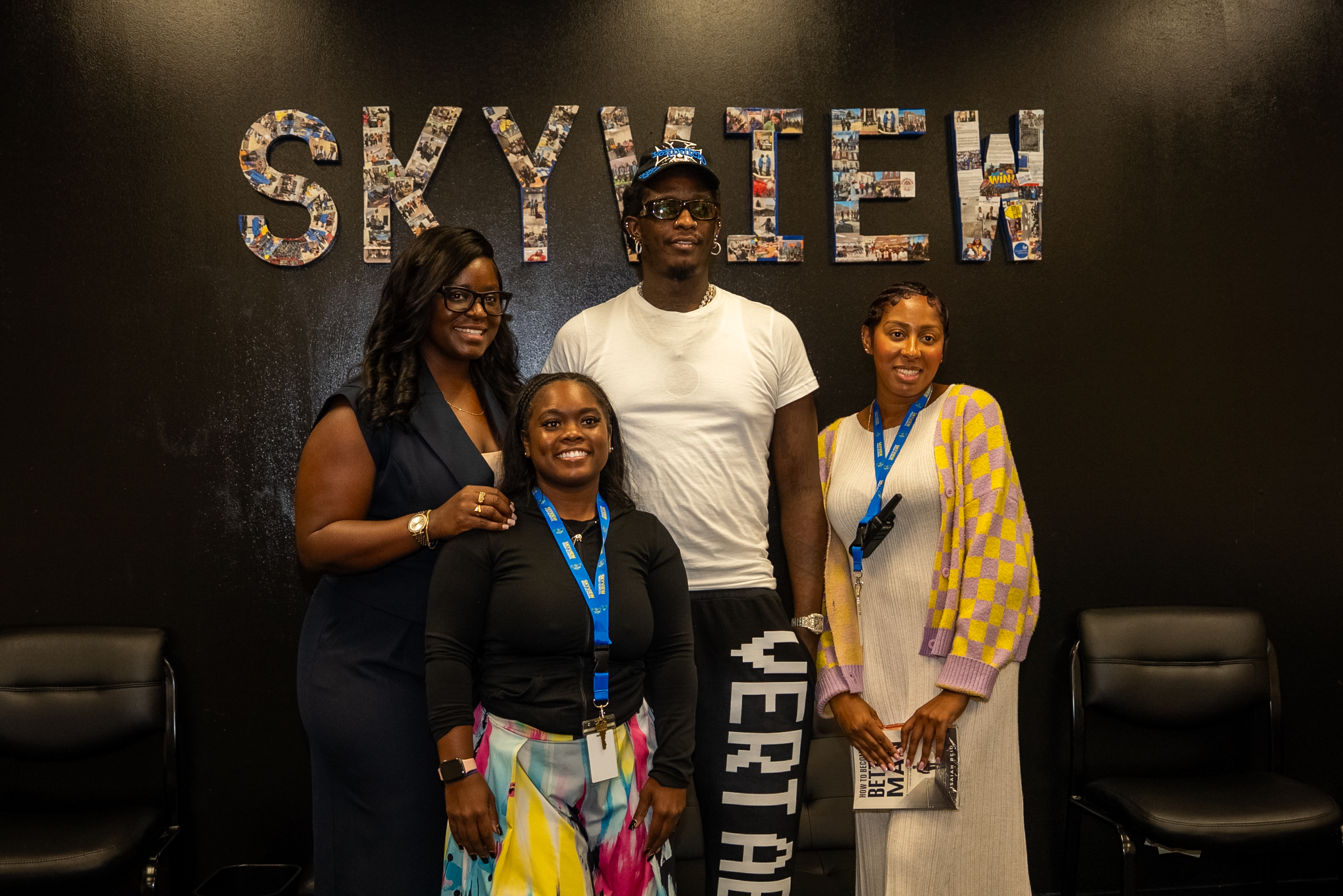 Four people standing in front of a large "SKYVIEW" sign, including a man wearing a white t-shirt and three women wearing colorful clothing.