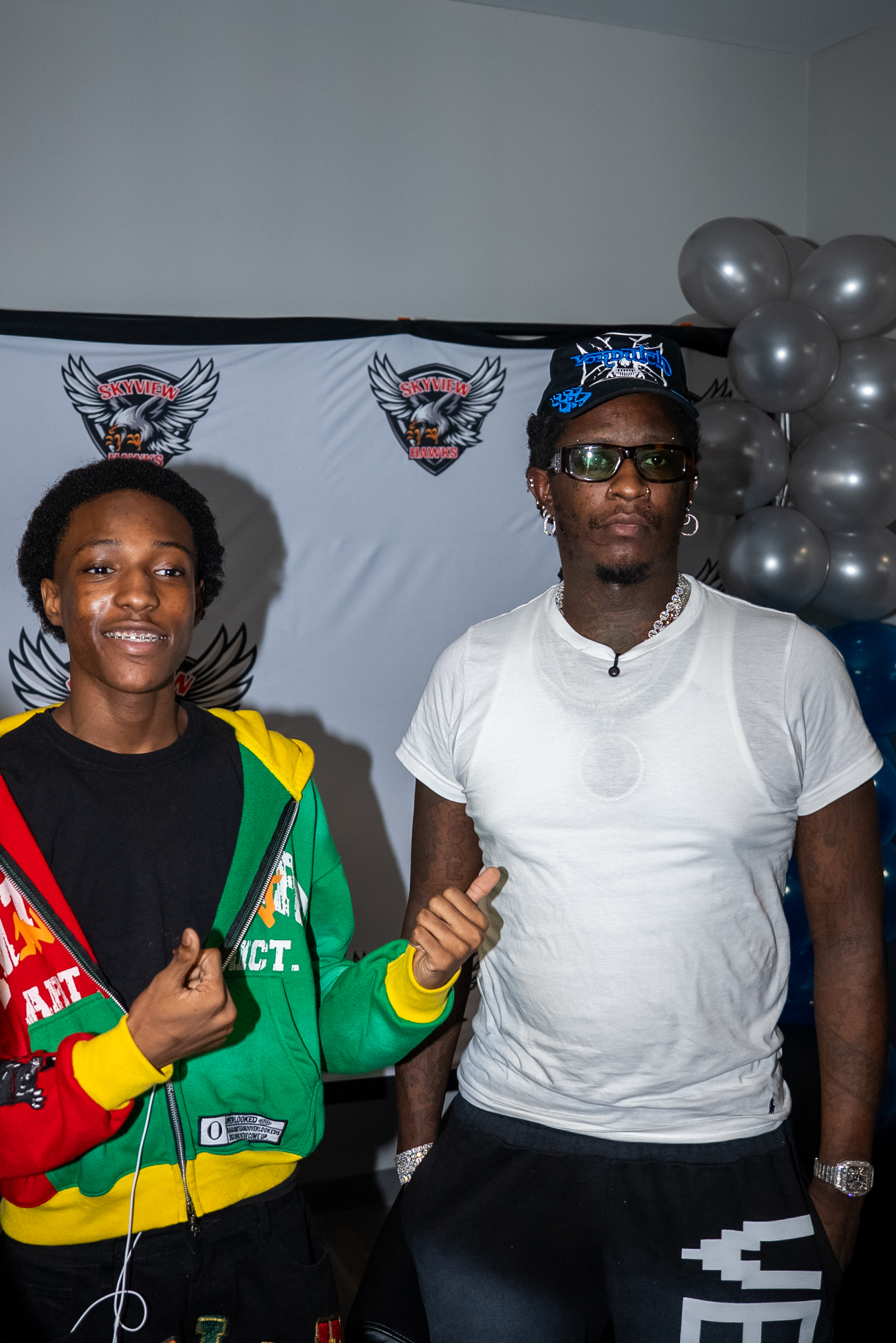 Two young men posing in front of a Skymile banner, one wearing a colorful jacket and the other in a white t-shirt and cap.