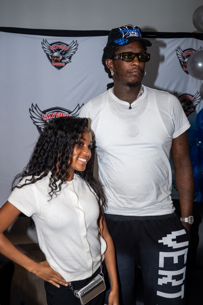 Two people, a man and a woman, standing in front of Skyview Hawks banners. The man is wearing a baseball cap, sunglasses, and a white t-shirt. The woman has curly hair and is smiling.
