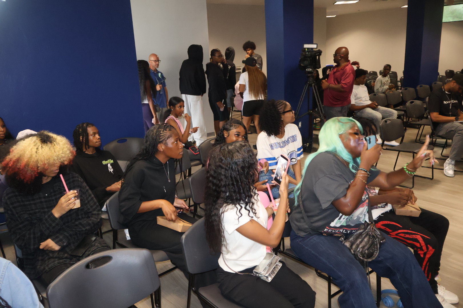 A group of people, mostly young adults, sitting in a room with cameras and equipment, suggesting a media or production event.