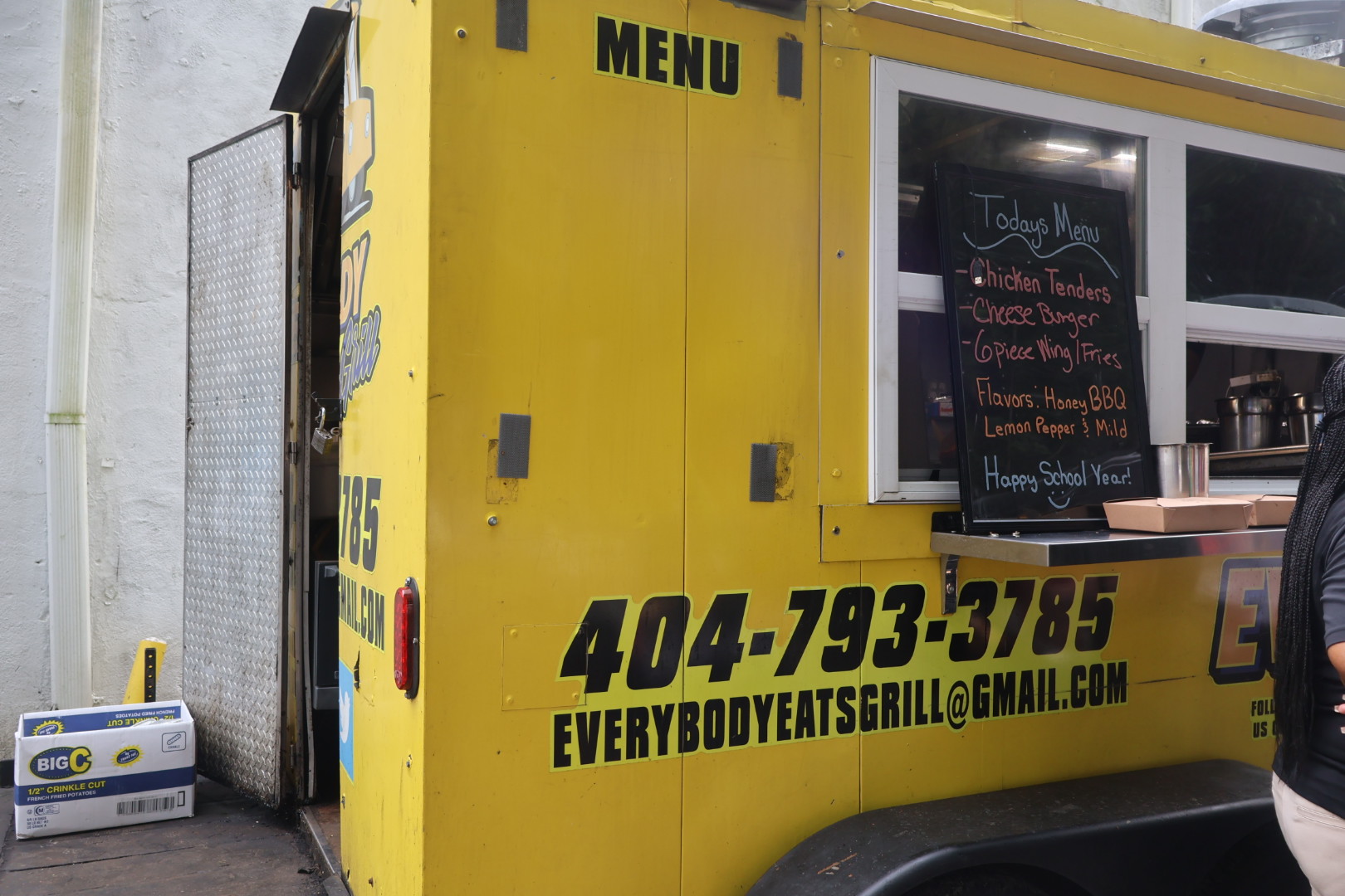 A yellow food truck with the text "MENU" and the phone number "404-793-3785" displayed prominently. The menu board lists items like "Chicken Tenders", "Cheese Burger", and "Waffle Fries".