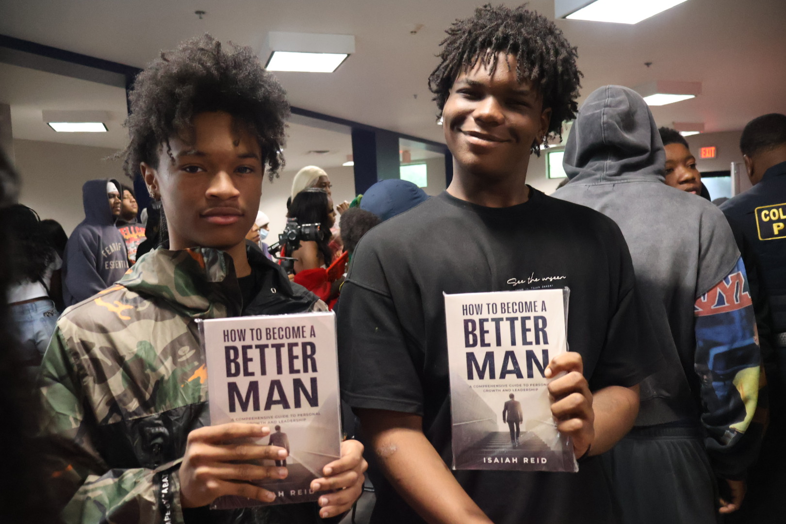 Two young men holding books titled "How to Become a Better Man" in a crowded indoor setting.