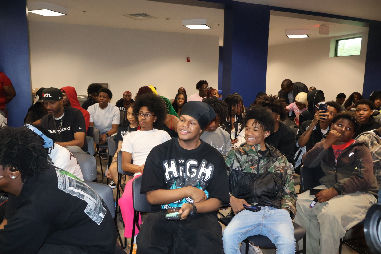 A crowded room of young people, many wearing t-shirts with the word "HIGH" printed on them, sitting and standing together.