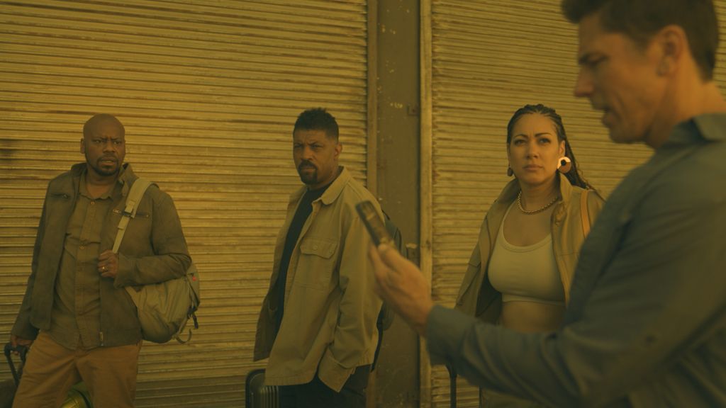 Four people standing in front of a closed storefront, one man holding a gun.