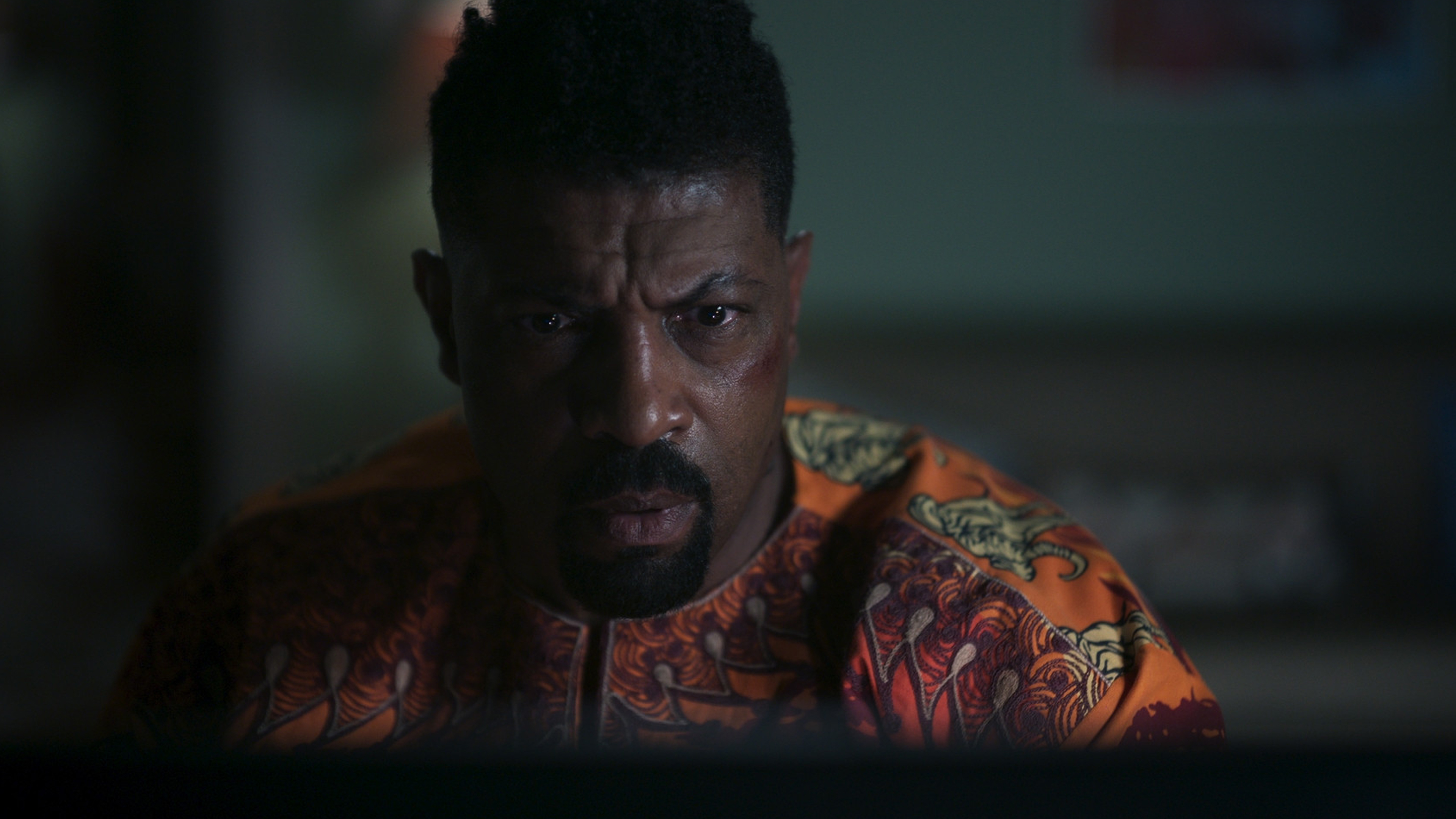 A close-up portrait of a serious-looking Black man with a beard, wearing an orange and black patterned shirt.