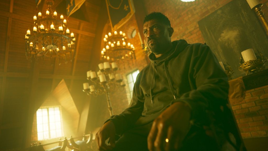 A man in a green robe sits in a dimly lit room with ornate chandeliers, gazing intently.