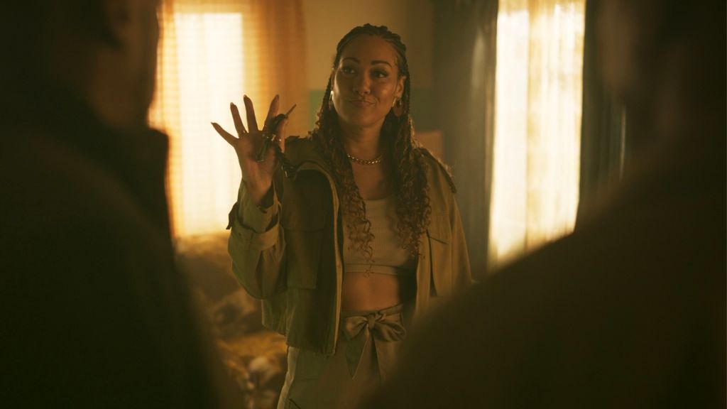 A woman with braided hair stands in a dimly lit room, her hand raised in a dramatic gesture.