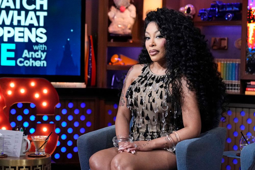 A woman with curly dark hair wearing a patterned dress sits on a couch in front of a "Watch What Happens Live with Andy Cohen" backdrop.