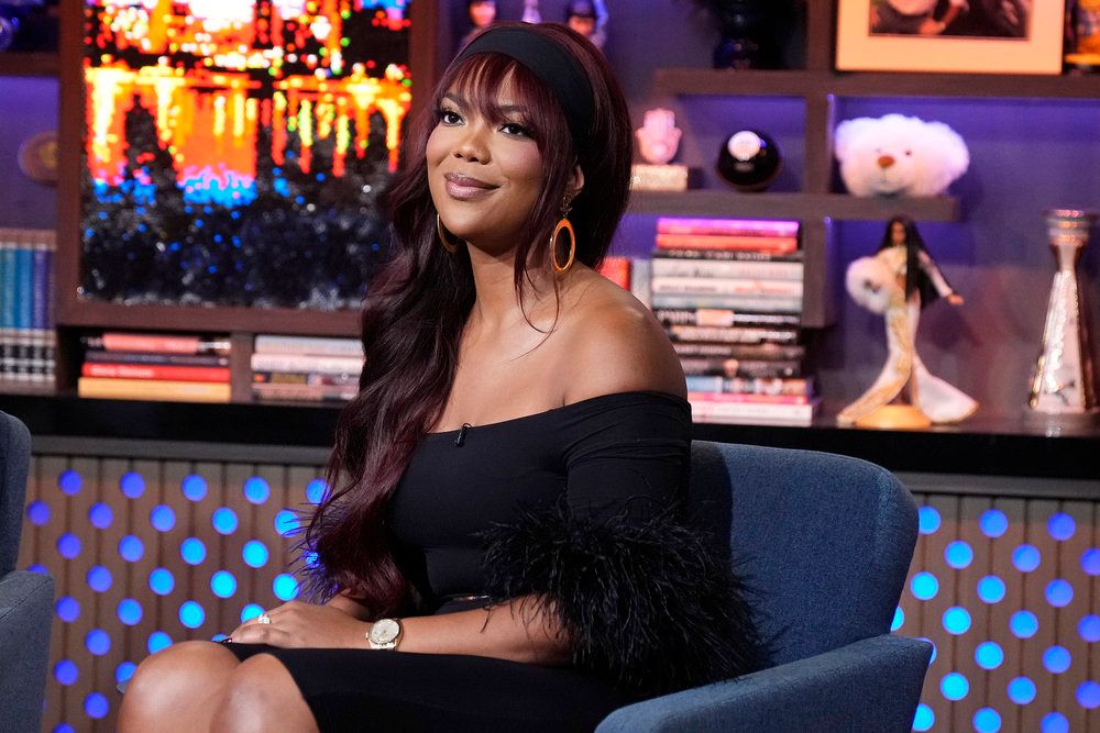 A woman with long dark hair wearing a black off-the-shoulder dress sits on a couch in a TV studio set with colorful lighting and shelves of books and decor.