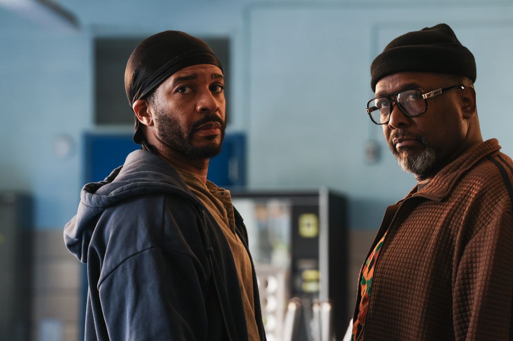 Two men, one wearing a black headband and the other glasses, standing in a room with blue walls.