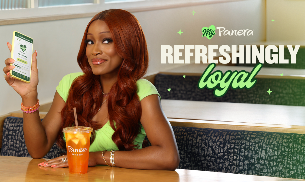 A woman with long red hair holding a Panera Bread drink and standing in front of a "My Panera Refreshingly Loyal" sign.
