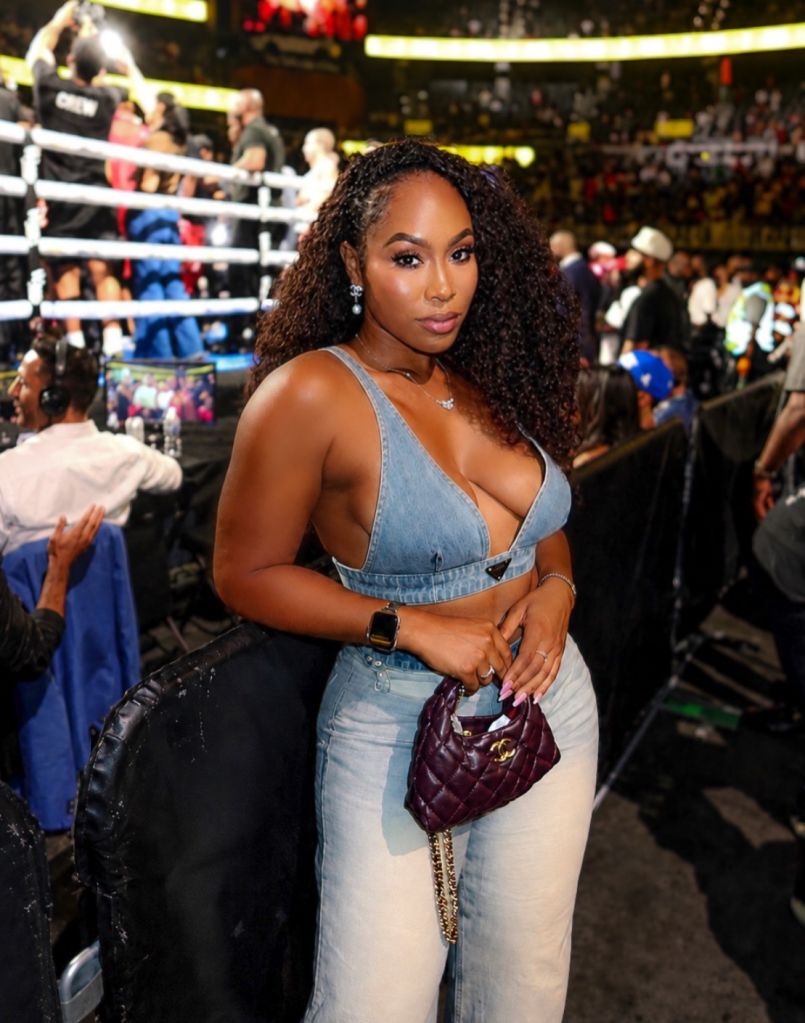 A woman with curly dark hair wearing a denim outfit and holding a burgundy handbag stands in front of a boxing ring.