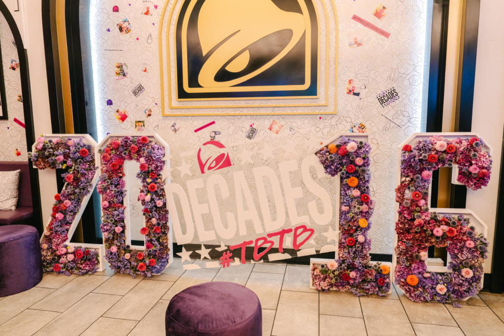 Decorative floral letters spelling "2023" against a backdrop of colorful flowers and a stylized eye logo, with a purple ottoman in the foreground.