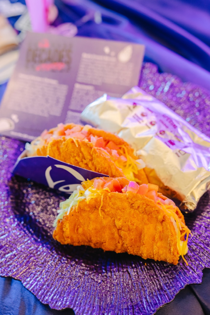 Closeup of two crispy fried tacos on a purple glittery surface, with a Taco Bell logo visible.