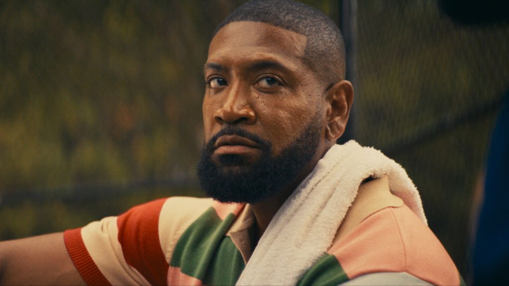 A close-up portrait of a Black man with a beard, wearing a colorful sweater and looking thoughtfully off-camera.