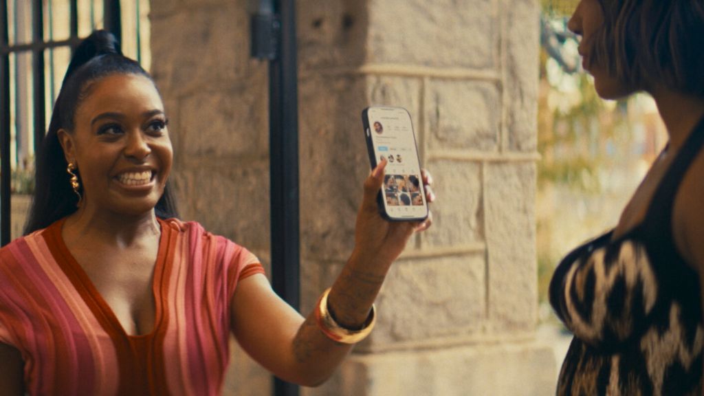 A smiling woman in a pink top holds a smartphone displaying a group photo on the screen, in front of a stone wall.