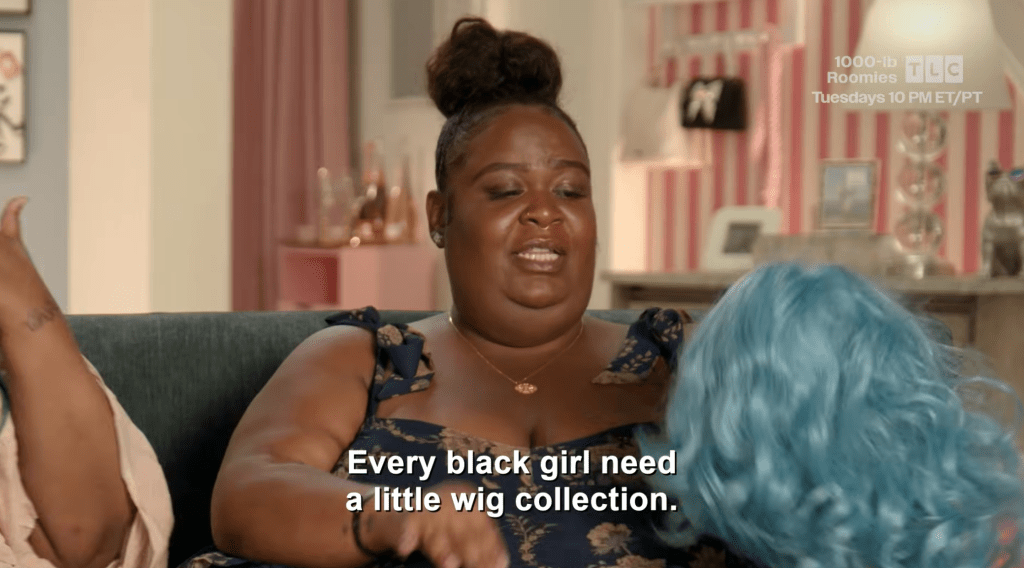A woman with a bun hairstyle wearing a patterned dress sitting on a couch and saying "Every black girl need a little wig collection.