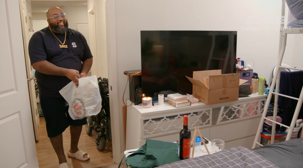 A smiling man with a beard wearing a black shirt and holding a plastic bag in a room with a TV, boxes, and other household items.