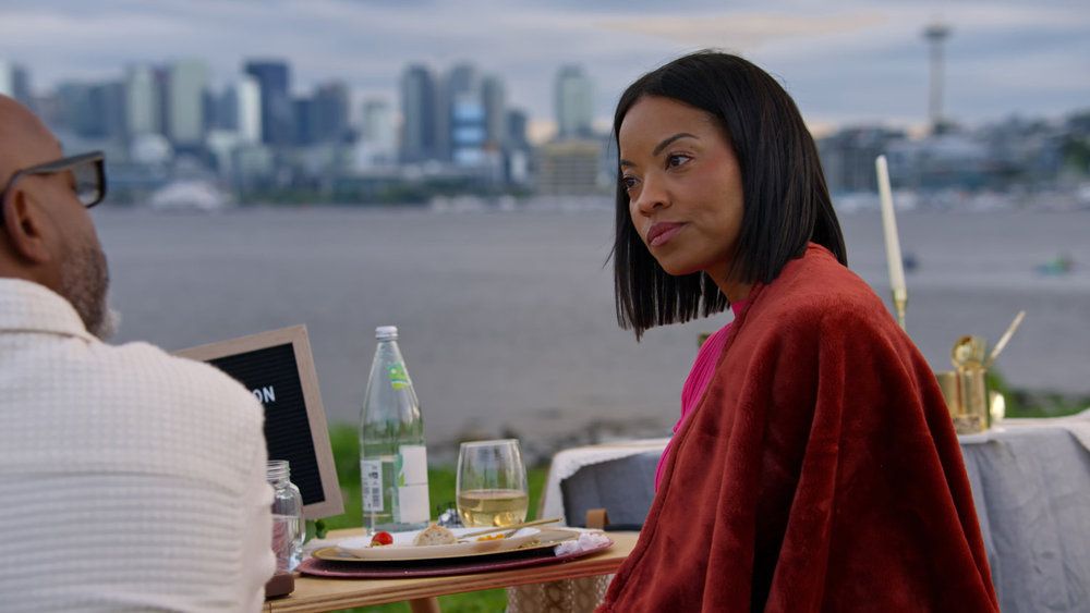 A woman in a red jacket stands on a balcony overlooking a city skyline, with a table and drinks in the foreground.