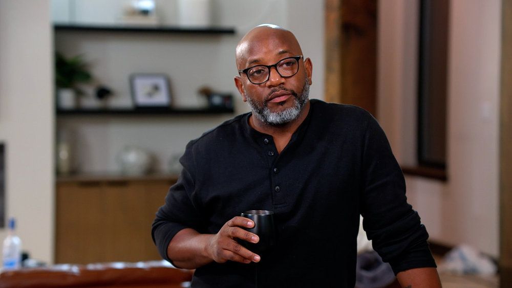 A middle-aged Black man with a beard and glasses, wearing a black shirt, standing in a room with shelves and decor.