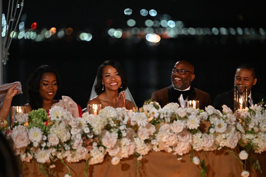 A wedding reception at night, with the bride, groom, and two other people smiling and surrounded by a floral centerpiece on a table.