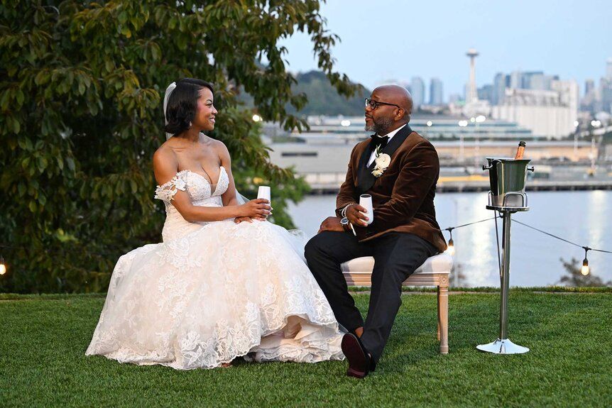 A bride and groom sitting on a bench in a garden setting, with a city skyline in the background and a champagne bucket on a stand next to them.