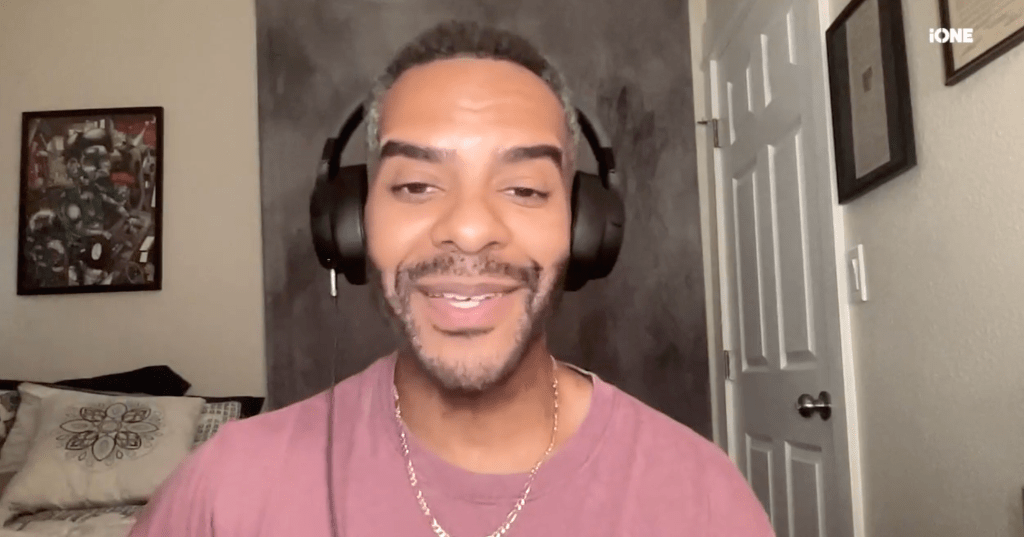 A smiling man wearing headphones and a pink shirt, sitting in a room with framed artwork on the walls.