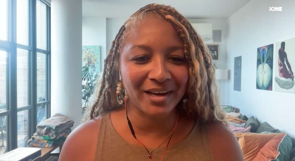 A smiling woman with braided hair wearing a tan top and jewelry, sitting in a room with artwork on the walls.
