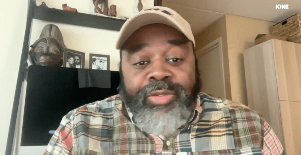 A middle-aged Black man with a beard wearing a baseball cap and plaid shirt, looking directly at the camera.