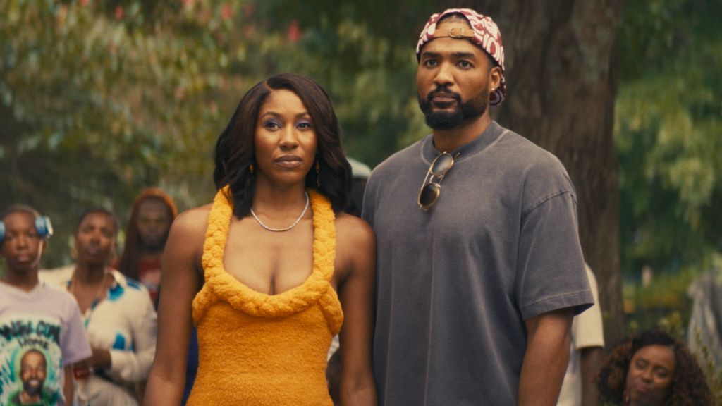 Two people standing together in an outdoor setting, a woman wearing a yellow top and a man wearing a gray shirt and headband.