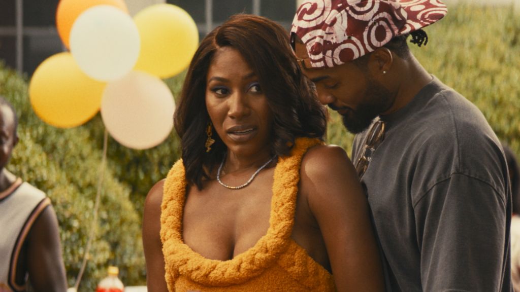 Two people standing in a field with balloons in the background. The woman is wearing a yellow furry top and the man is wearing a patterned hat.