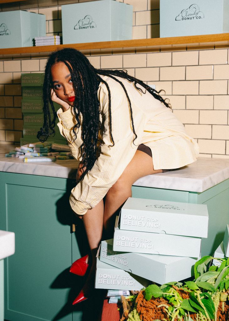A person with long dreadlocks wearing a white jacket and red heels, sitting on a counter surrounded by 